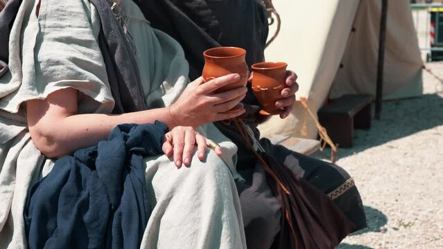 Medieval couple in linen clothing sitting on wooden bench in legionaries camp and drinking wine from clay mugs, man hugging woman while talking, everyday life of ancient Roman people in military camp