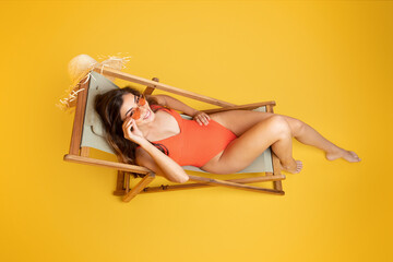 Cheerful millennial asian woman in swimsuit and sunglasses sit on deck chair, enjoy free time