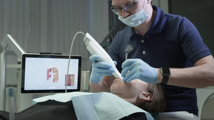 Doctor scans the patient's teeth in the clinic. The dentist holds in his hand a manual 3D scanner for the jaw and mouth. Dental health. Creates a 3D model of teeth and gums on a medical monitor - obrazy, fototapety, plakaty