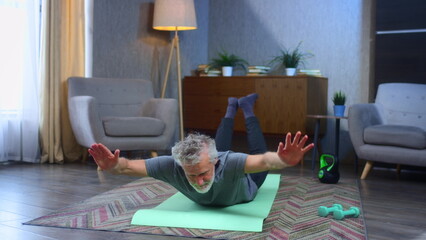 Portrait of a gray-haired senior man with a beard. Doing exercises on fitness mat. Grandfather working out at home in living room. An old man in sportswear makes a plank for the back