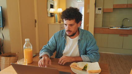 Fototapeta na wymiar Guy dressed in denim shirt works on laptop while having lunch in cozy kitchen