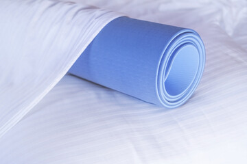 A blue rolled up yoga fitness mat is rolled up on a bed, lying on a pillow under a blanket,  Yoga fitness morning practice concept