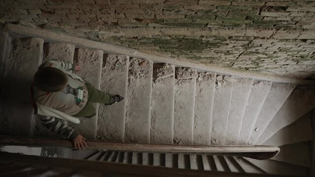 Lonely caucasian boy descends an abandoned dusty staircase in a house, top view. Old wooden steps in a crumbling building. Many traces of people from shoes on a dirty surface.