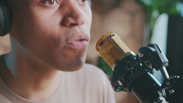 Close up mouth of young African American man near condenser microphone for recording radio broadcast or podcast for internet listeners sits in home recording studio. Journalist, speaker