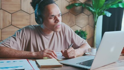 Young attentive African American man in headphones participates in online meeting and writes down tasks of project manager in notebook sits at table with papers and laptop. Job, career, freelance
