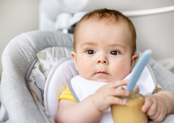 adorable cute baby boy sitting in swing cradle eating mashed food with spoon.mother hand feeding infant toddler.child with bib is smiling,no teeth on gum.vegetables or fruits puree in jar.hungry