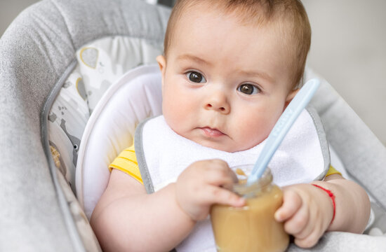 adorable cute baby boy sitting in swing cradle eating mashed food with spoon.mother hand feeding infant toddler.child with bib is smiling,no teeth on gum.vegetables or fruits puree in jar.hungry