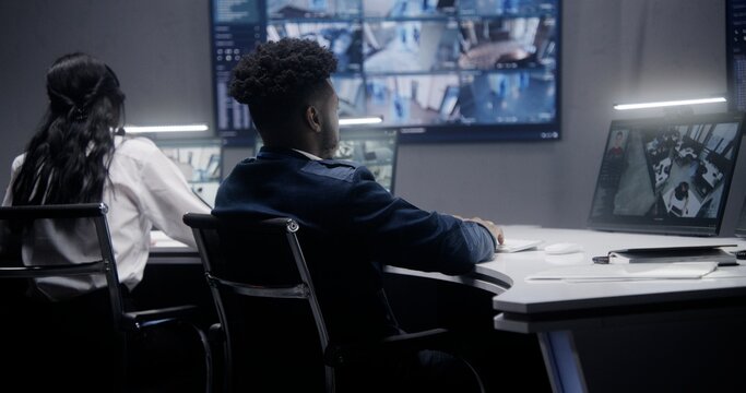 African American security operator monitors CCTV cameras with AI face recognition. Multiple big screens on the wall showing security cameras view. Colleague works at background. Social safety concept.