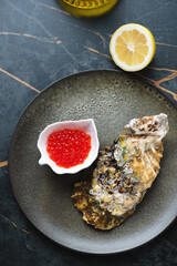 Olive-colored plate with unopened oyster and red caviar, vertical shot on a dark-olive marble background, above view