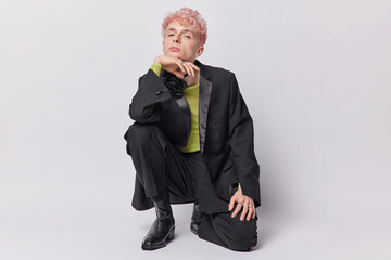 Self assured adult man has serious expression squats against white background keeps hand under chin...