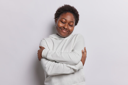 Body positive young woman embraces herself has good self esteem wears casual hoodie has eyes closed dreams about something isolated over white background. Body acceptance and self love concept