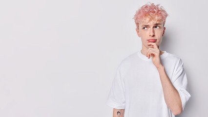 Displeased sad man with pink hair keeps hand on chin looks unhappily aside dressed in casual t shirt planss about something isolated over white background copy space for your advertising content