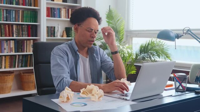 Young tired African American man student looks into laptop and takes off glasses scratches head suffering from error in test for online university sits at table with crumpled papers in home office.