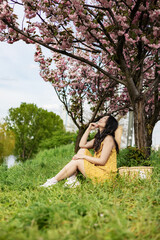 Naklejka na ściany i meble Happy young Asian woman in a wicker hat walks in a blooming city park. Portrait of a stylish emotional model in a yellow dress near pink flowering trees.
