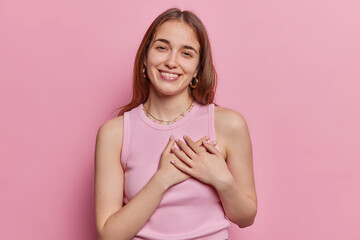 Tender cheerful European woman dark straight hair shows grateful gesture presses hands to heart smiles pleasantly dressed in casual t shirt isolated over pink background expresses happiness.