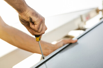 The detail of an unrecognizable technical specialist hand is installing a hot water solar panel with a screwdriver on a roof of a residential. Concept of solar panels, renewable energy.