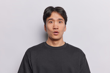Surprised young Asian man with dark short hair feels amazed stands speechless reacts to shocking news dressed in casual black t shirt poses against white background. Human reactions concept.