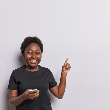 Vertical shot of dark skinned woman smiles toothily messages with smartphone uses mobile phone app points index finger at copy space dressed in casual black t shirt isolated over white background