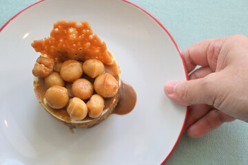 Hand holding plate of delicious toffee macadamia nut cake serving customers. Small round homemade toffee cake topped with macadamia nuts. Topped with sweet caramel and chocolate slices.popular dessert