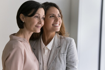 Middle-aged mom and young adult, grown up daughter wear elegant clothes posing standing indoors, daydreaming staring out window. Family bond and understanding, two colleagues at workplace. Friendship