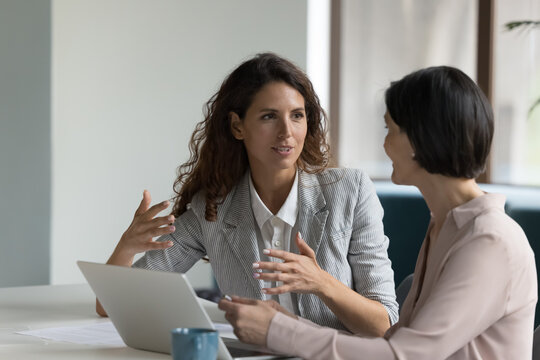 Two business women sit at desk discuss project details, diverse female colleagues met in office, share opinion, working on collaborative task, sales manager makes commercial offer to company client
