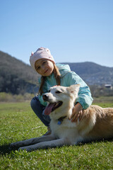 Children and pets concept. Kid is smiling, sitting in a clearing and stroking a white domestic dog of mixed breed. Charming little Caucasian girl walks in park with her dog.