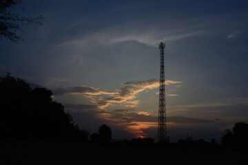 A tall radio tower silhouetted against a colorful sunset sky. There are trees in the foreground.