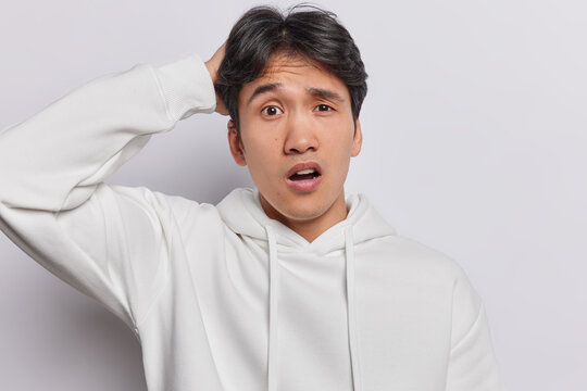 Confused dark haired Asian man looks embarrassed keeps mouth widely opened stares with puzzlement scratches head dressed in casual hoodie isolated over white background. Human face expressions