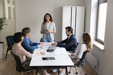 Group of cheerful businesspeople gather in conference room during morning briefing, take part in formal meeting, joking having friendly workflow in modern office, training led by Hispanic female coach