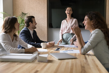 Group of successful businesspeople gathered in conference room, distracted from briefing, laughing, looking happy, engaged in corporate meeting event or training led by mature female coach. Business