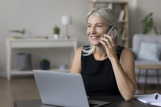 Cheerful happy middle aged entrepreneur woman talking on cellphone at table with laptop, using wireless online connection technology for work from home, smiling, enjoying mobile phone conversation