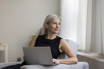 Thoughtful positive blonde mature woman resting in armchair, holding laptop, typing, looking at window away, thinking, dreaming. Senior freelancer lady working at home