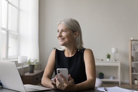 Happy dreamy freelancer, entrepreneur woman sitting at work table, using smartphone, desktop computer, looking away, smiling, laughing, enjoying wireless communication technology at home