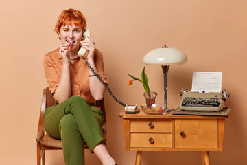 Cheerful retro redhead female writer makes phone call via stationary telephone sits on chair near table with vintage typewriter and lamp wears old fashionable clothes isolated over brown background