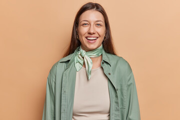 Portrait of attractive dark haired woman smiles happily wears green shirt and kerchief around neck being in good mood poses against brown background feels very positive. People and emotions concept