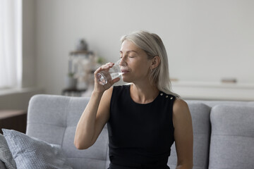 Peaceful pretty blonde mature woman drinking natural mineral water with closed eyes for healthcare, healthy metabolism, refreshment, sitting on couch at home, satisfying thirst - obrazy, fototapety, plakaty