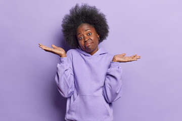 Confused dark skinned young woman with bushy hair spreads palms with hesitation shrugs shoulders...