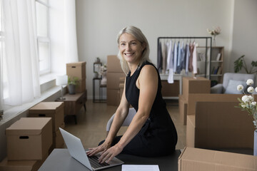 Cheerful senior entrepreneur woman typing on laptop at home storage place, leaning at work table, looking at camera, smiling, laughing, Internet shop owner portrait with stacked boxes in background