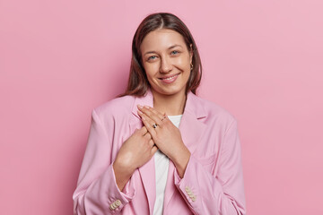 Pretty dark haired woman presses hands to chest smiles gently dressed in formal jacket looks directly at camera expresses gratitude poses against pink studio background. You belong to my heart