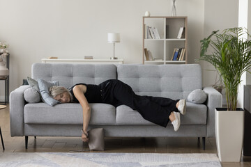 Exhausted sleepy mature woman in casual clothes and shoes holding hand bag, resting on couch, lying...