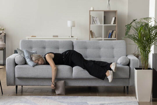 Exhausted sleepy mature woman in casual clothes and shoes holding hand bag, resting on couch, lying on belly with closed eyes, falling asleep, taking break, pause, feeling fatigue, burnout