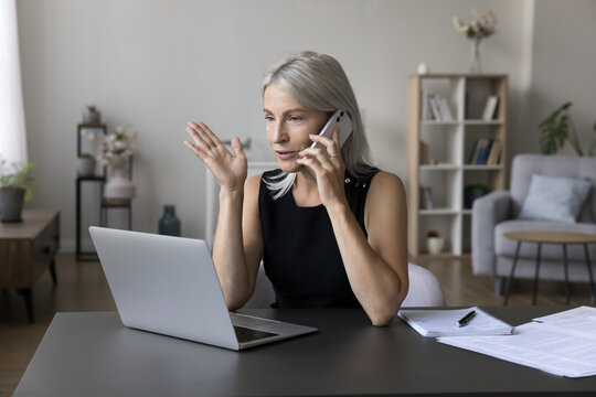 Positive busy mature freelance business woman speaking on mobile phone at home workplace, sitting at laptop, making call, speaking, using digital gadgets for work from home