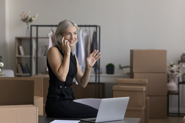 Positive middle aged internet shop owner working in storage space, talking on cellphone at laptop...