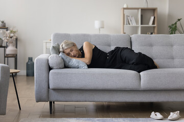 Calm middle aged blonde homeowner woman lying on side on comfortable sofa, sleeping at daytime, feeling tired, exhausted, fatigue, relaxing, enjoying domestic leisure time