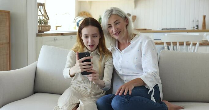 Cheerful teenage kid girl and mature blonde grandma talking on video call, holding smartphone, looking a screen ,speaking, laughing, grimacing, having fun, resting on home couch