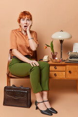 Vertical shot of shocked retro woman with short red hair exclaims loudly keep mouth widely opened dressed in old fashioned clothing gets important task to do poses on chair with vintage bag on floor