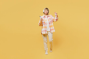 Full body elderly blonde caucasian woman 50s years old she wear casual clothes headphones listen to music use mobile cell phone isolated on plain yellow background studio portrait. Lifestyle concept.