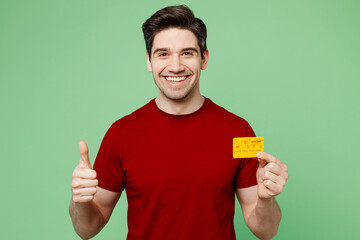 Young smiling happy man he wearing red t-shirt casual clothes hold in hand mock up of credit bank card show thumb up isolated on plain pastel light green background studio portrait. Lifestyle concept.