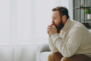 Side view young minded caucasian ginger man wearing casual clothes look aside sitting on grey sofa couch stay at home hotel flat rest relax spend free spare time in living room indoor. Lounge concept.