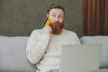 Young ginger man wears casual clothes use laptop pc computer hold credit bank card shopping online sit on grey sofa couch stay home hotel flat rest relax spend free spare time in living room indoor.